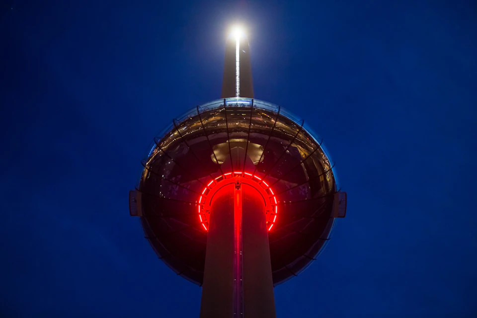 A glowing observation tower at night with a brightly illuminated circular pod and a light at the top, set against a dark sky.