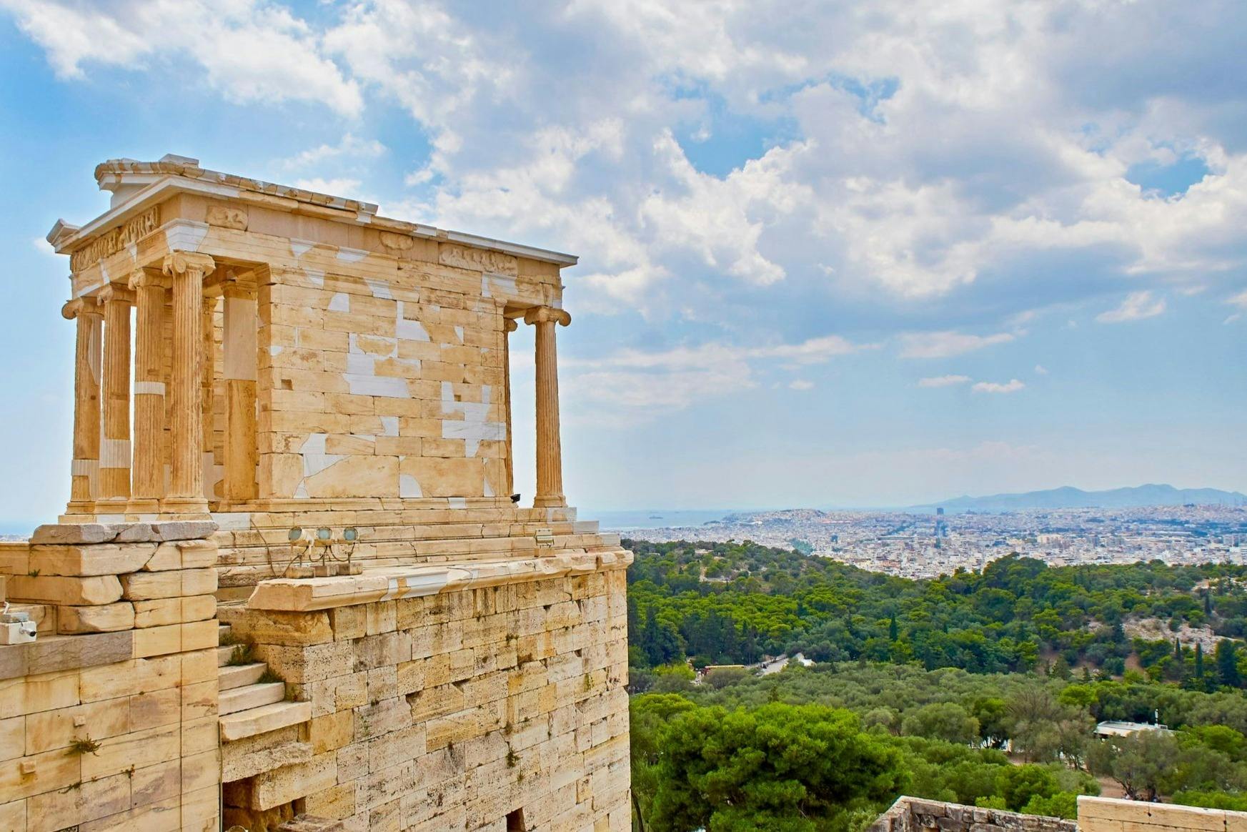 Αρχαίο πέτρινο κτίριο με κίονες με θέα ένα καταπράσινο τοπίο και μια μακρινή πόλη κάτω από έναν εν μέρει συννεφιασμένο ουρανό.