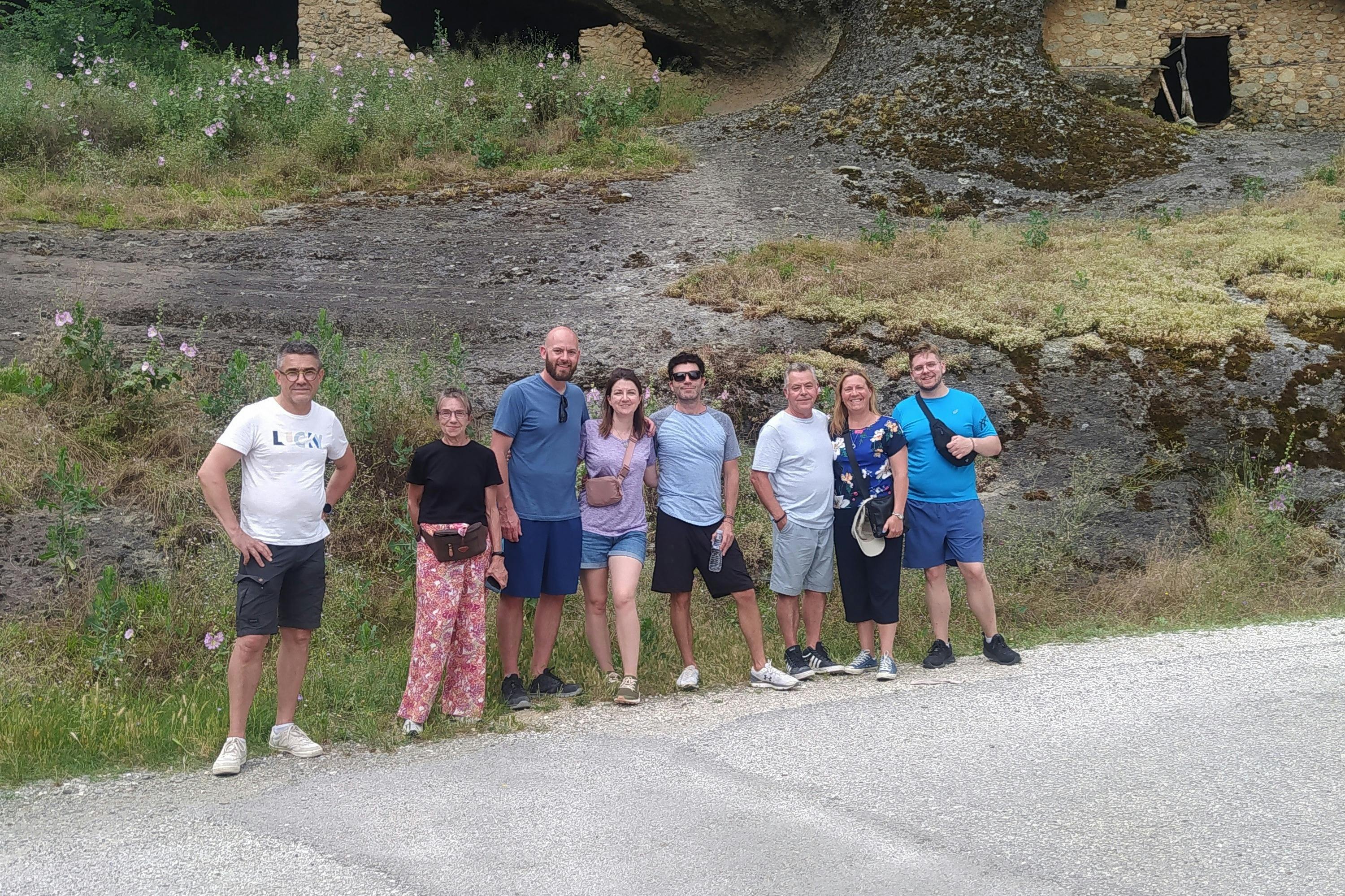 Een groep van zeven mensen staat buiten op een weg bij een heuvel met vegetatie en ruïnes op de achtergrond.