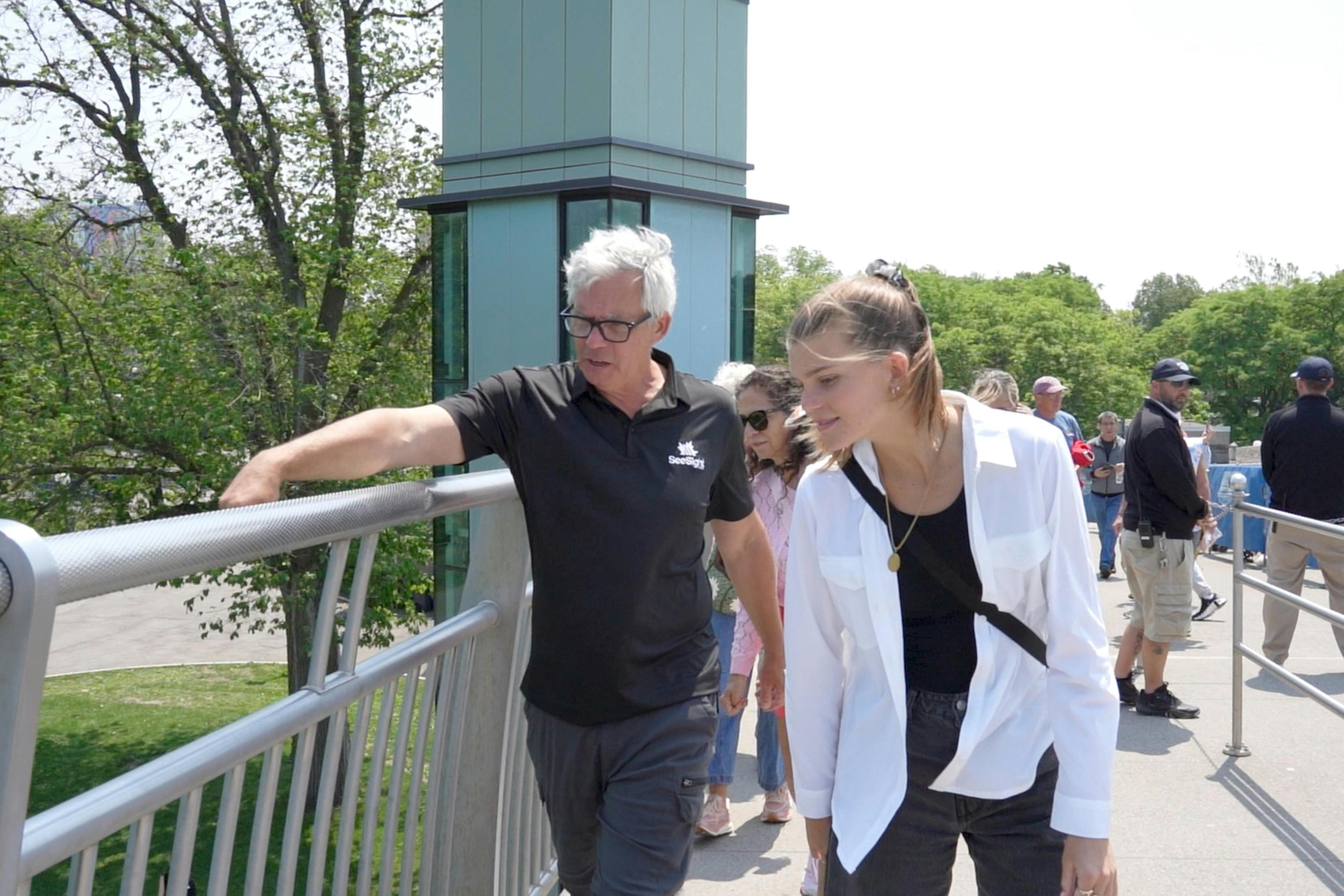 검은색 셔츠를 입은 남성과 흰색 셔츠를 입은 여성을 비롯해 다리를 걷고 있는 사람들로, 푸른 나무가 배경을 이루고 있습니다.