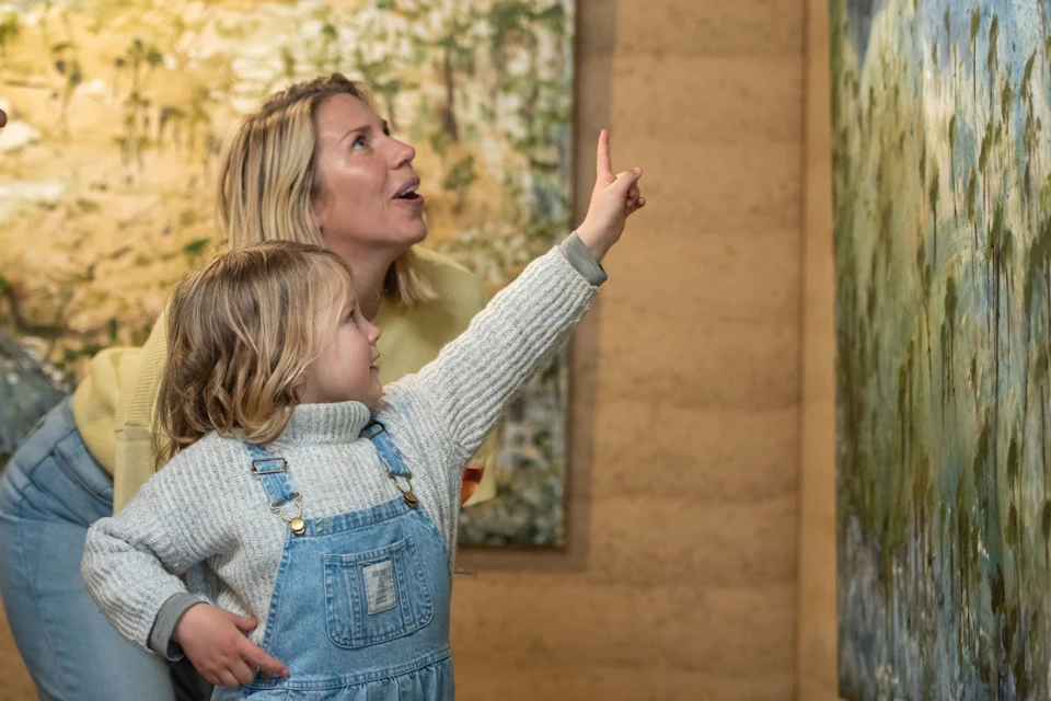 A woman and a young child point and look up while indoors, with a blurred painting in the background.
