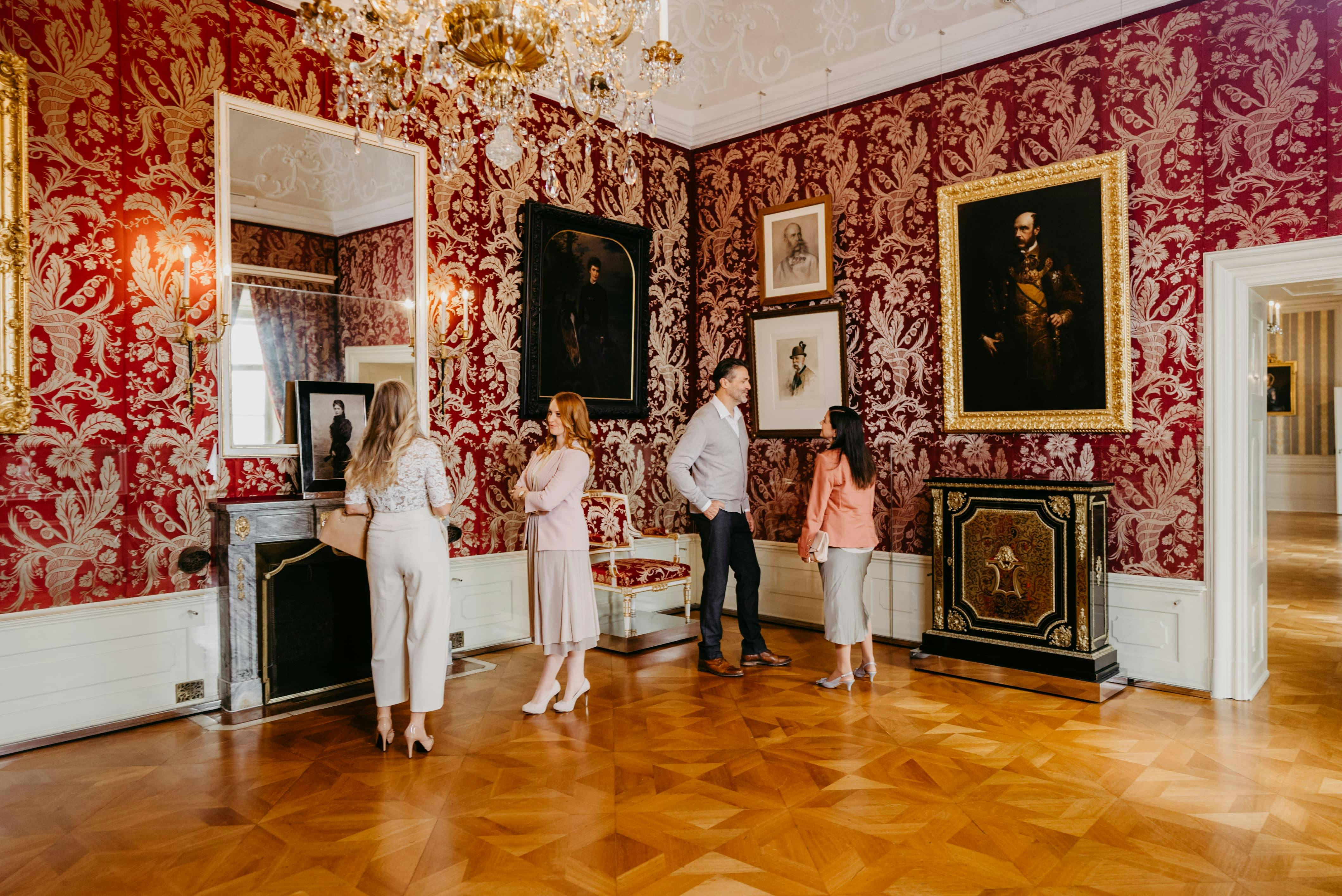 Άνθρωποι σε ένα πολυτελές δωμάτιο με κόκκινη ταπετσαρία δαμασκηνών, περίτεχνο πολυέλαιο και κορνιζαρισμένα πορτρέτα στους τοίχους.