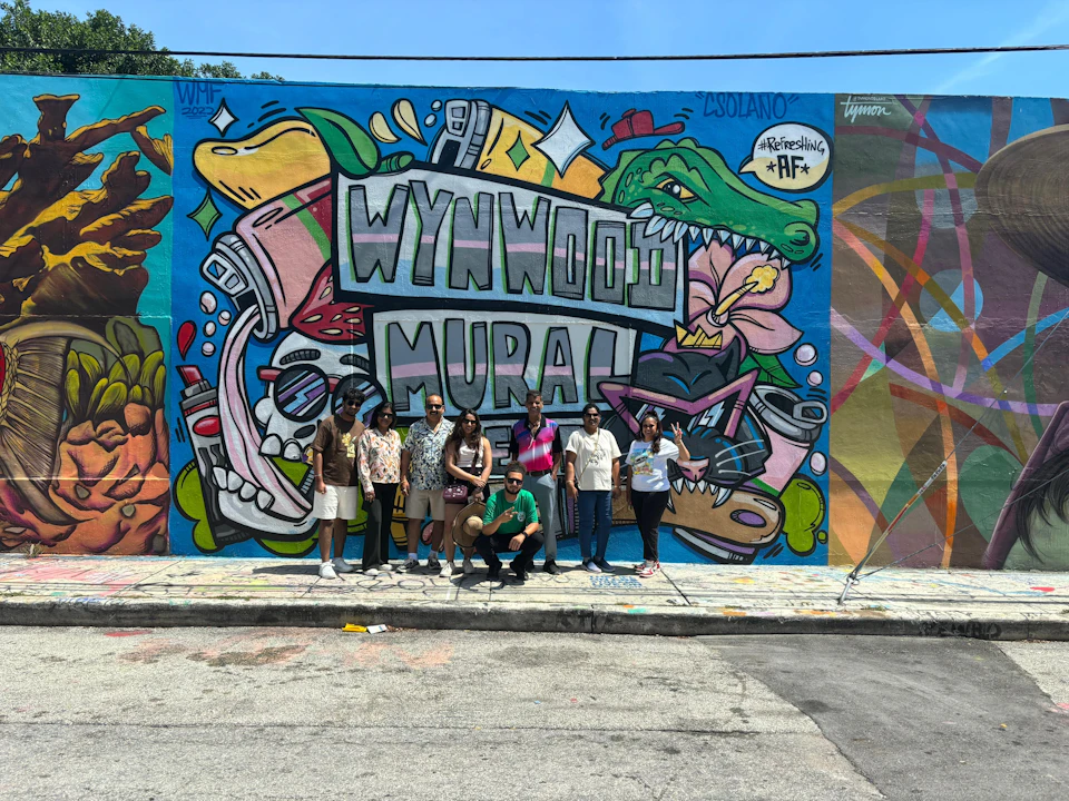 A group of people posing in front of a colorful mural with the text "Wynwood Mural" and various vibrant illustrations.