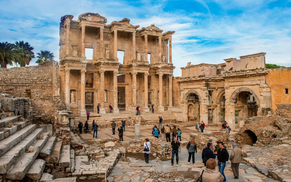 Tourists explore the ancient ruins of the Library of Celsus in Ephesus, with its grand façade and marble columns under a blue sky.