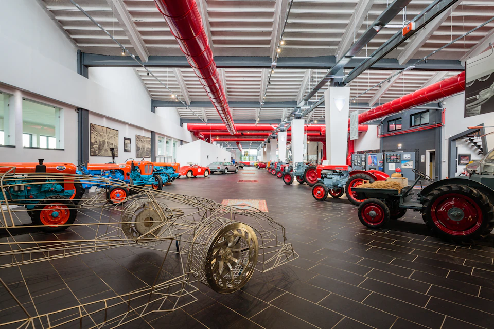 A spacious indoor exhibit showcasing vintage tractors and a wireframe car model, with red ceiling pipes and large windows.