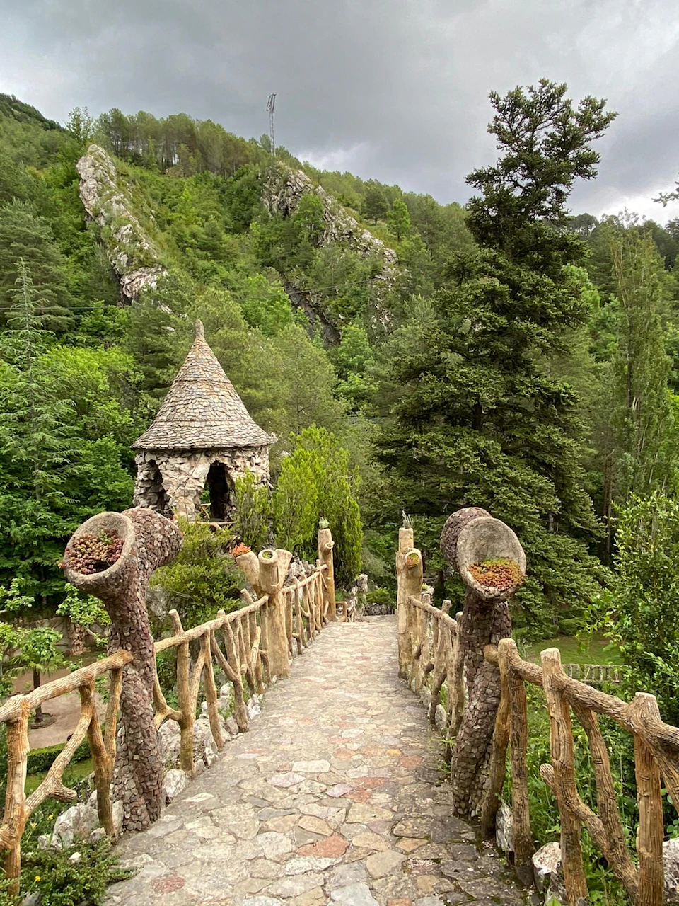 A stone pathway with a rustic wooden bridge leads to a small, round stone hut with a conical roof, surrounded by dense greenery.