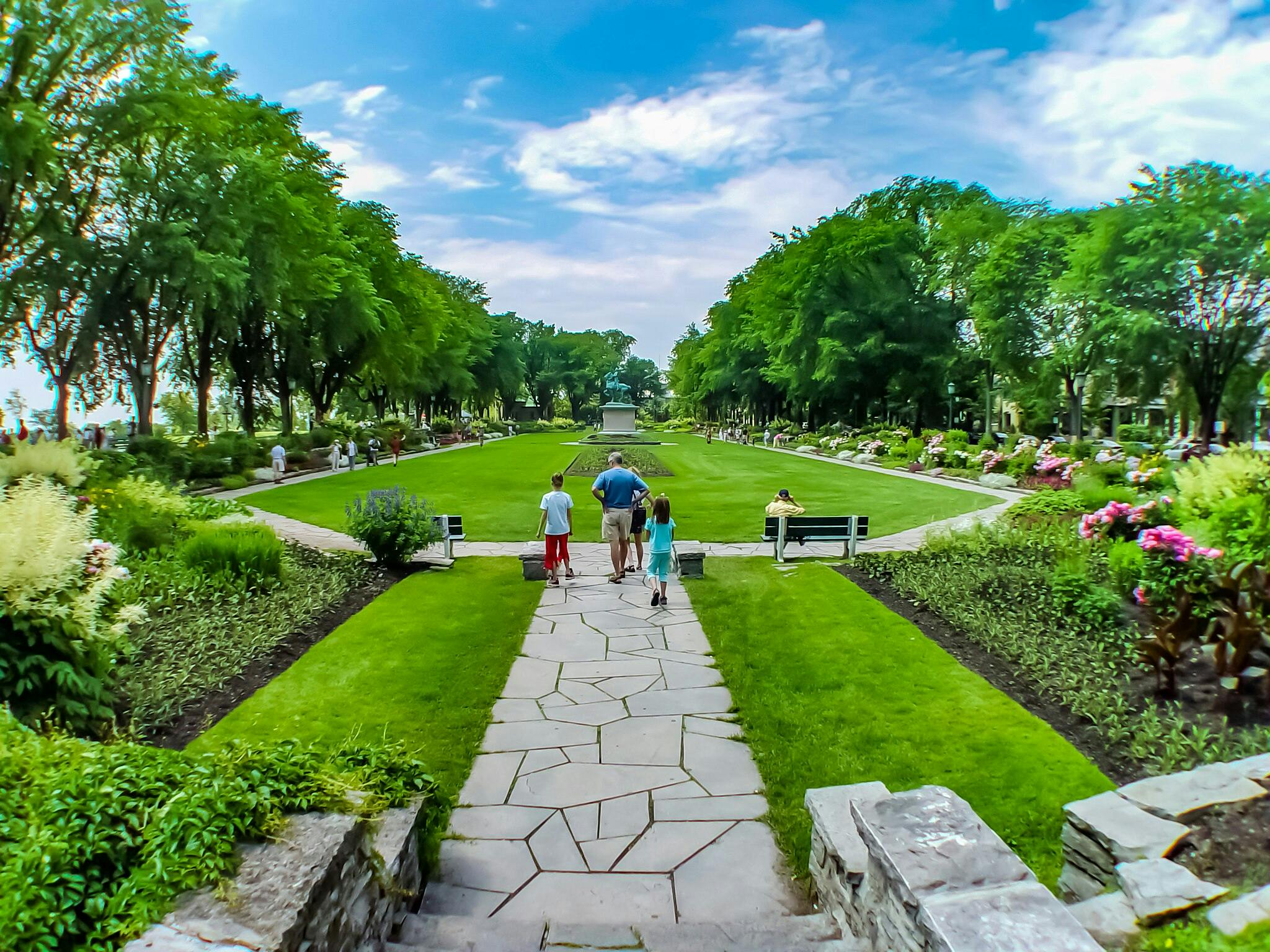 People walking on a pathway in a park with lush gardens, benches, and tall trees under a partly cloudy sky.