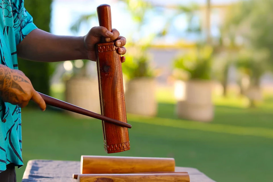A person holding a wooden percussion instrument outdoors, tapping it with a stick, with blurred greenery in the background.
