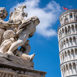 En marmorstaty av tre keruber ligger i förgrunden med det lutande tornet i Pisa synligt mot en blå himmel i bakgrunden.