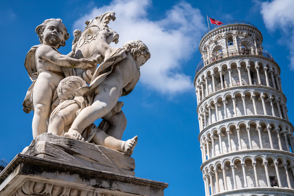 A marble statue of three cherubs is in the foreground with the Leaning Tower of Pisa visible against a blue sky in the background.