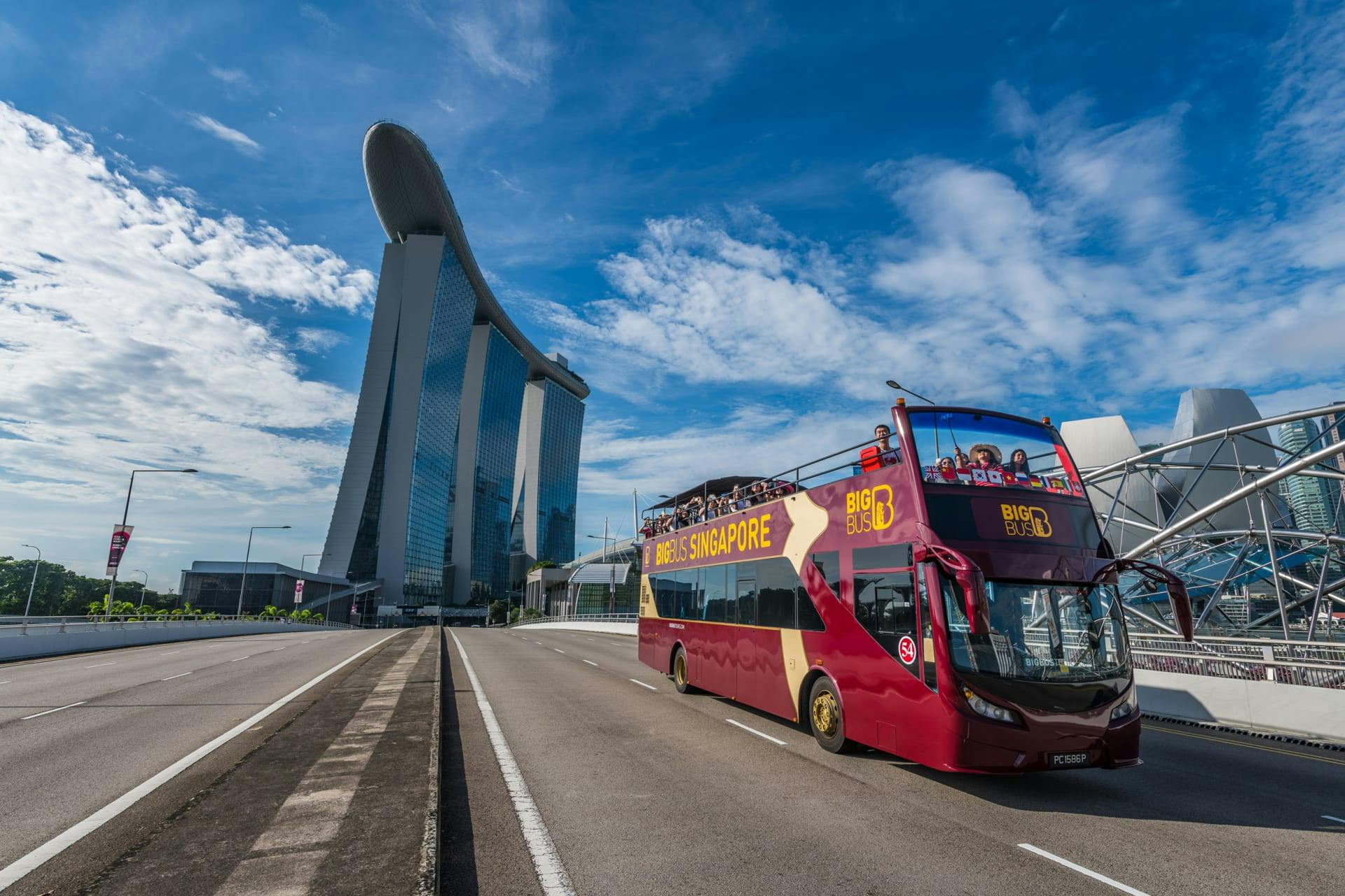 Un bus touristique à impériale passe devant un grand bâtiment moderne sous un ciel bleu clair parsemé de nuages.