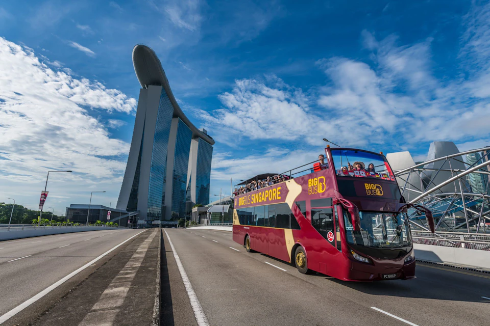 A double-decker tour bus drives past a tall, modern building under a clear blue sky with scattered clouds.