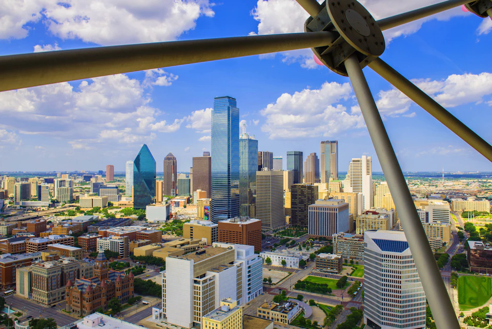 A skyline view of a city with tall buildings, a blue sky with white clouds, and a metallic structure in the foreground.