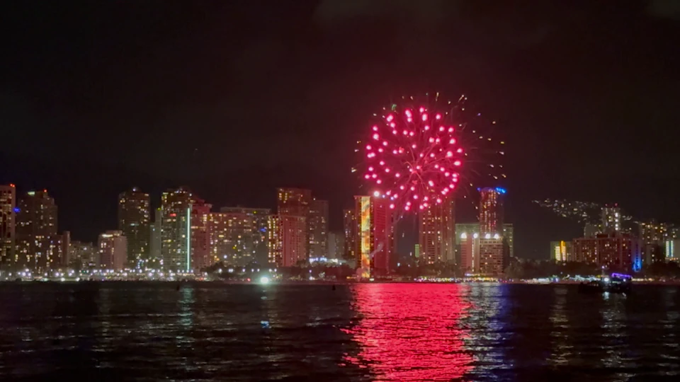 Bright red fireworks burst in the night sky above a city skyline, their reflection visible on the water below.