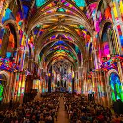 En katedralinteriör med livfull mångfärgad belysning på väggar och tak, fylld med en sittande publik.