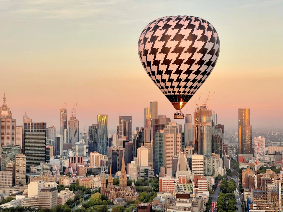 A hot air balloon with a black and white houndstooth pattern floats above a cityscape featuring various high-rise buildings at sunset.