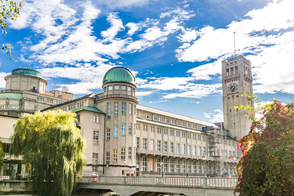 A large historic building with a green dome and clock tower under a bright blue sky, with a tree and bridge in the foreground.