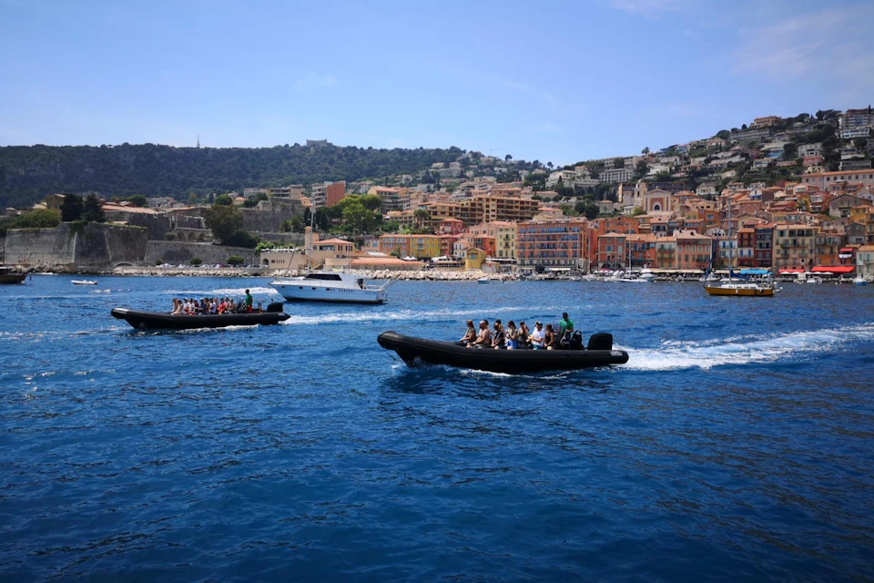 People riding inflatable boats on a blue sea near a colorful coastal town with hills in the background.