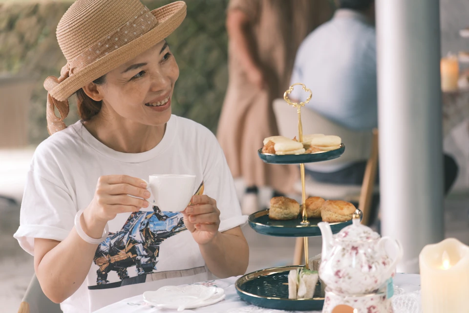 A woman wearing a straw hat sits at a table holding a teacup, with a tiered tray of pastries in front of her.