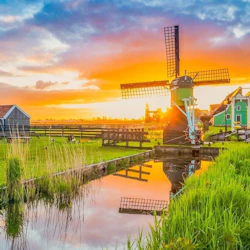 A windmill near a small canal with grassy surroundings and a colorful sunset sky in the background.