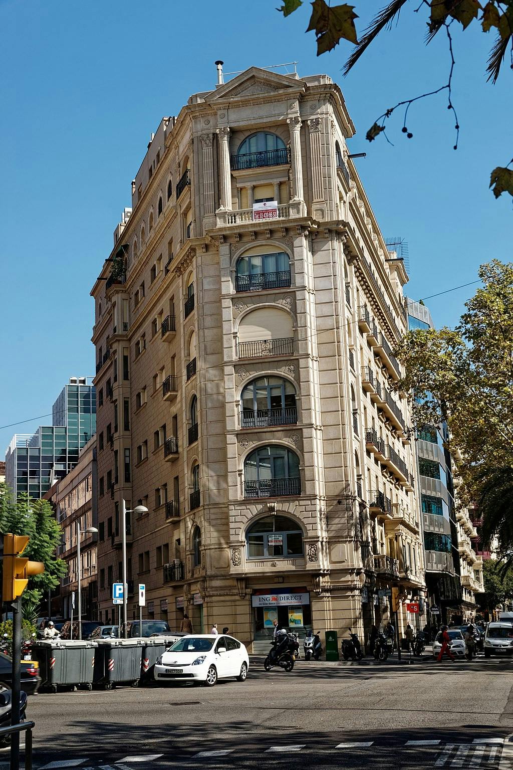 Γωνία δρόμου με ένα μπεζ, πολυώροφο κτίριο με καμάρες και μπαλκόνια. Αυτοκίνητα, μοτοσικλέτες και πεζοί που φαίνονται παρακάτω.