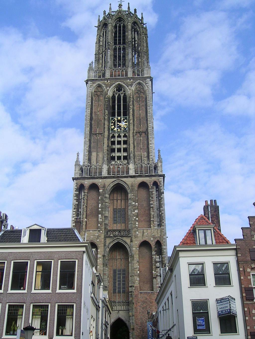 Tall brick and stone gothic tower with arched windows and a clock, flanked by buildings under a partly cloudy sky.