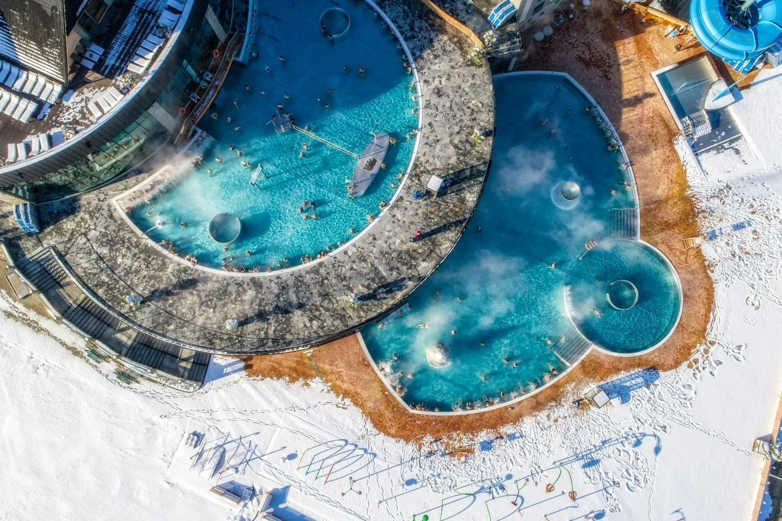 Widok z lotu ptaka na odkryte baseny z unoszącą się parą, otoczone śniegiem. Ludzie relaksują się w basenach i leżą na leżakach.