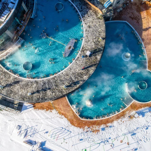 Flygfoto över utomhuspooler med ånga som stiger, omgiven av snö. Folk kopplar av i poolerna och ligger på solstolar.