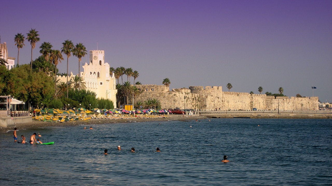 People swimming near a beach with colorful umbrellas, palm trees, a white building, and an ancient stone wall under a purple sky.
