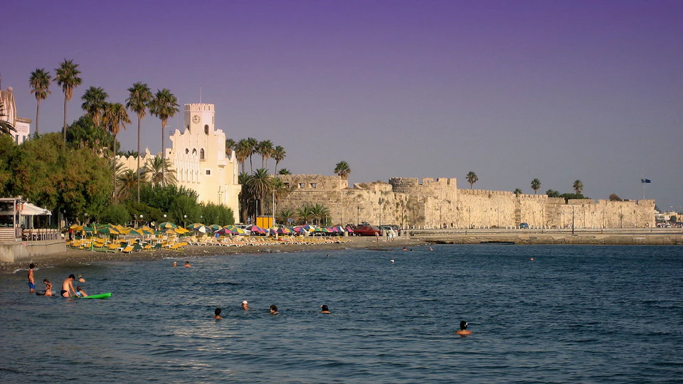 People swimming near a beach with colorful umbrellas, palm trees, a white building, and an ancient stone wall under a purple sky.