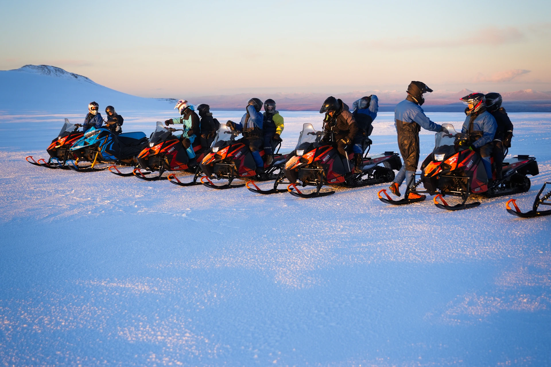 A group of people riding snowmobiles on a snowy plain under a clear sky during sunset.