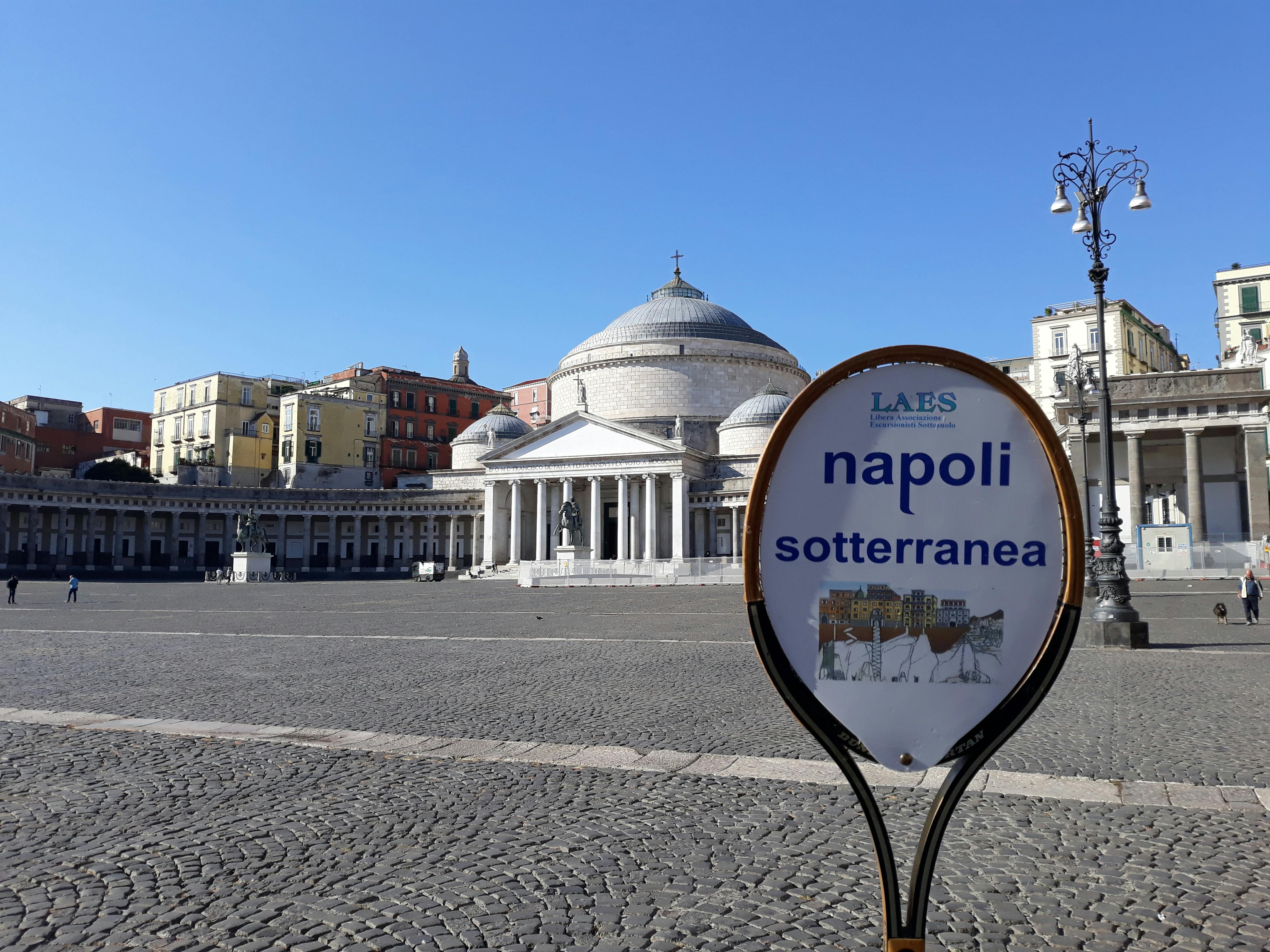 「那不勒斯Sotterranea」的标志位于一个大型开放式广场的前景，后面是一座圆顶建筑和五颜六色的建筑。