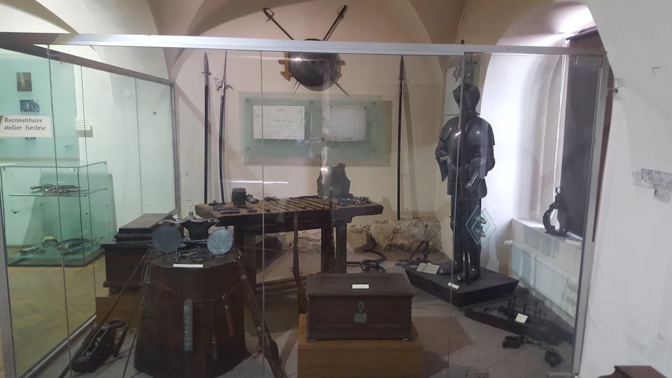 Museum exhibit showcasing historical armor, weapons, a wooden chest, and documents. A mannequin in armor stands on the right.