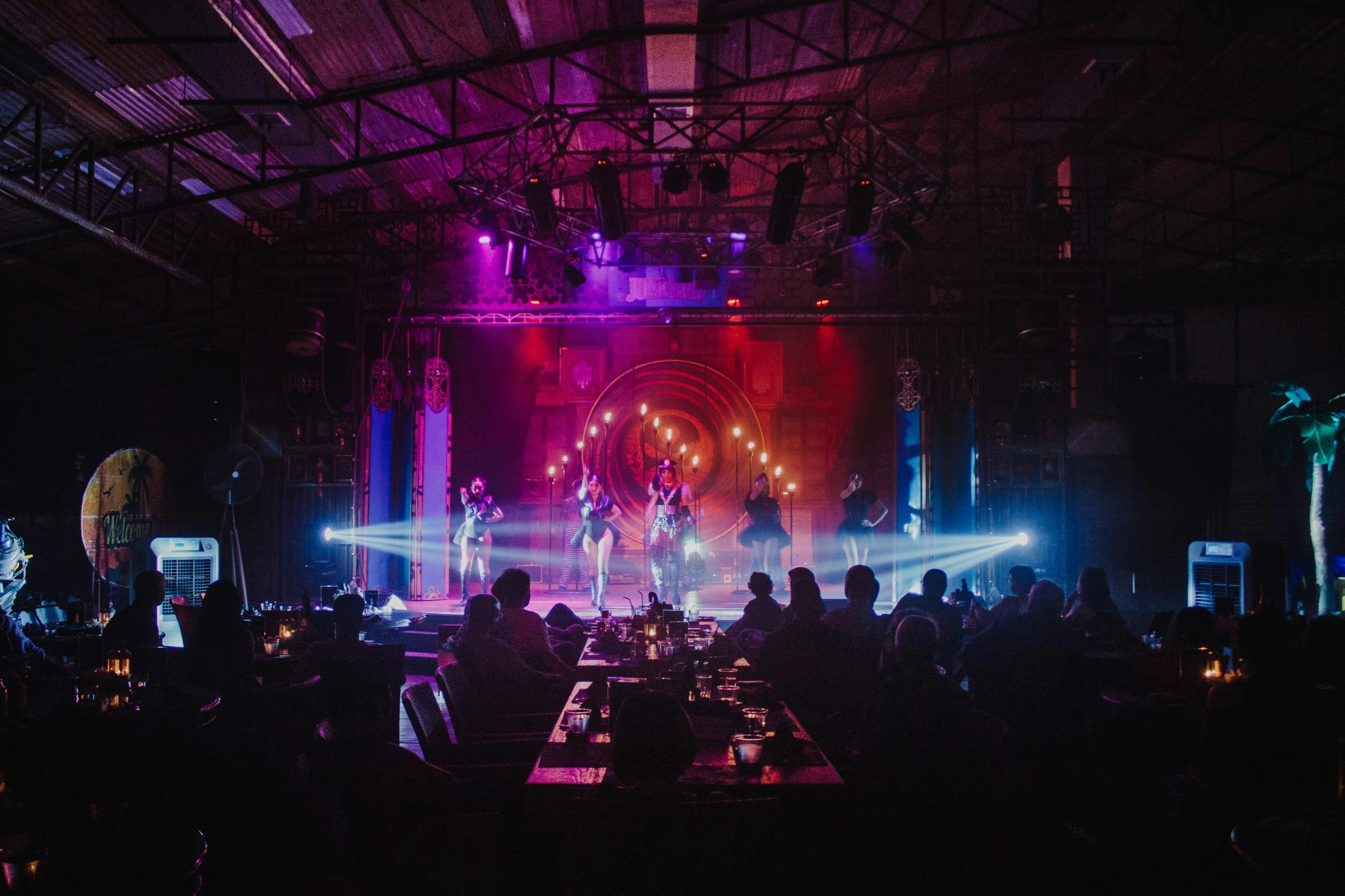 A stage performance with colorful lights and dancers in a venue with an audience seated at tables in the foreground.