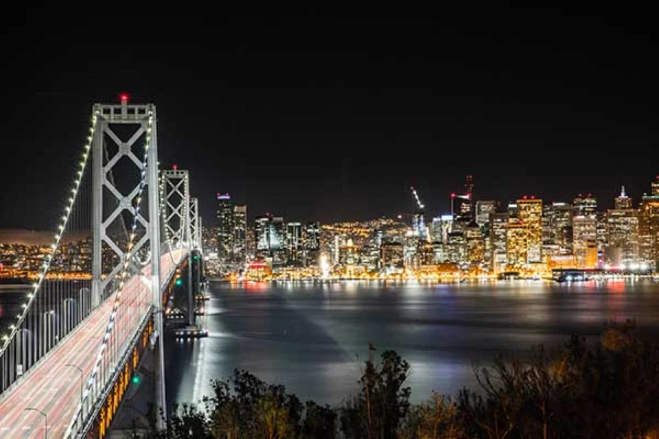 Night view of a city skyline with illuminated skyscrapers and a brightly lit suspension bridge over a calm body of water.