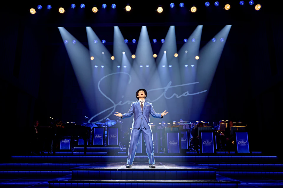 A performer in a blue suit and hat sings passionately on a stage under spotlights, with a band and "Sinatra" script behind.