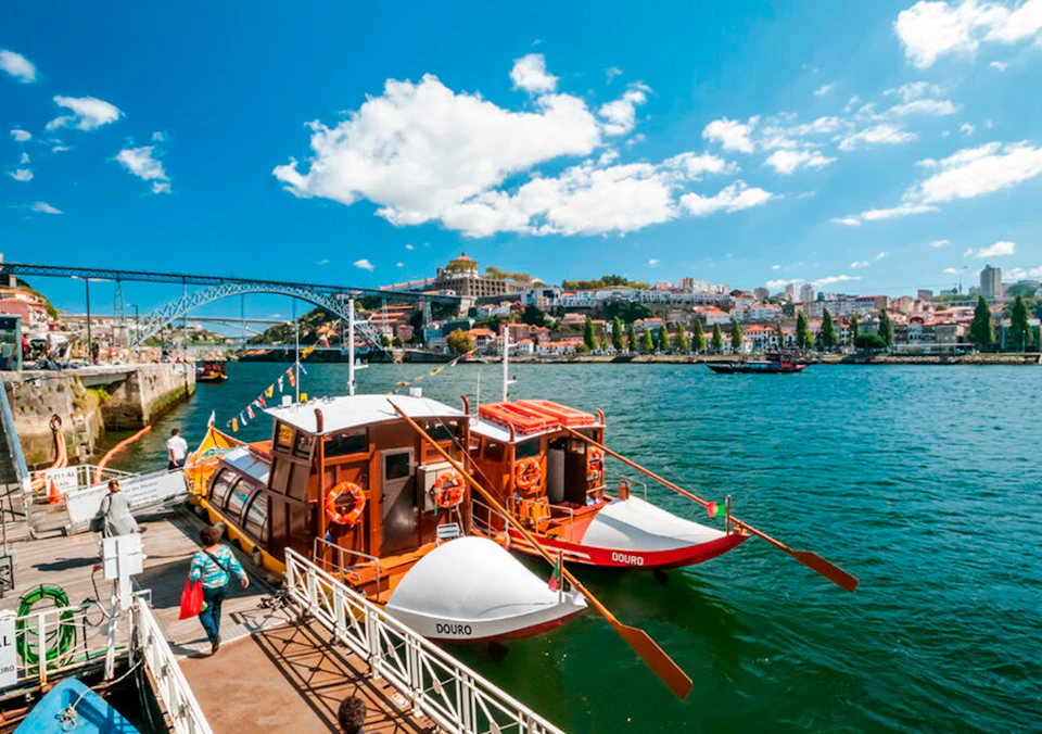 Two colorful boats docked on a river, with a cityscape, bridge, and people walking on the pier under a blue sky with clouds.