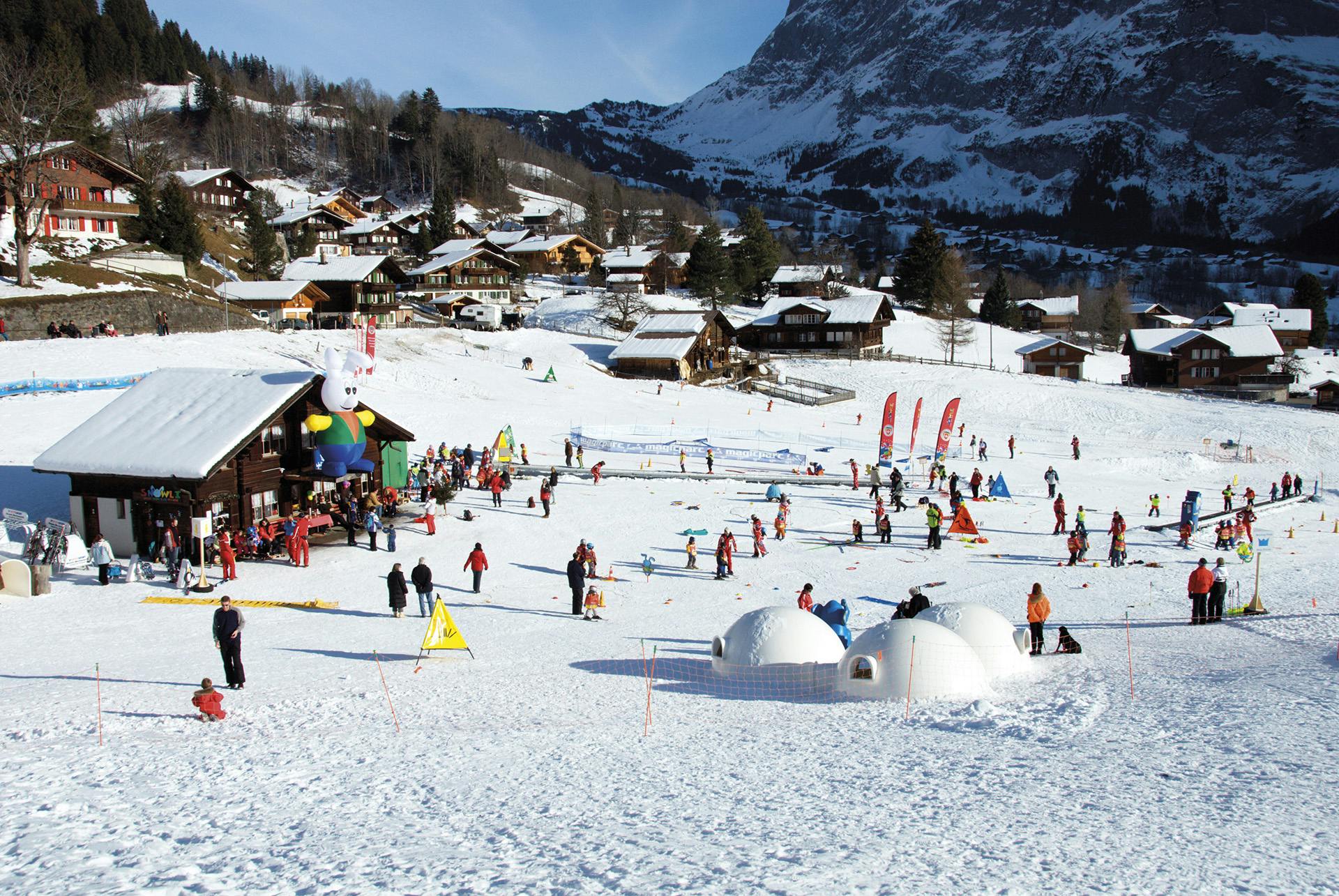 야외 활동을 즐기는 사람들이 있는 눈 덮인 마을. 산을 배경으로 이글루, 화려한 깃발, 나무 오두막이 보입니다.
