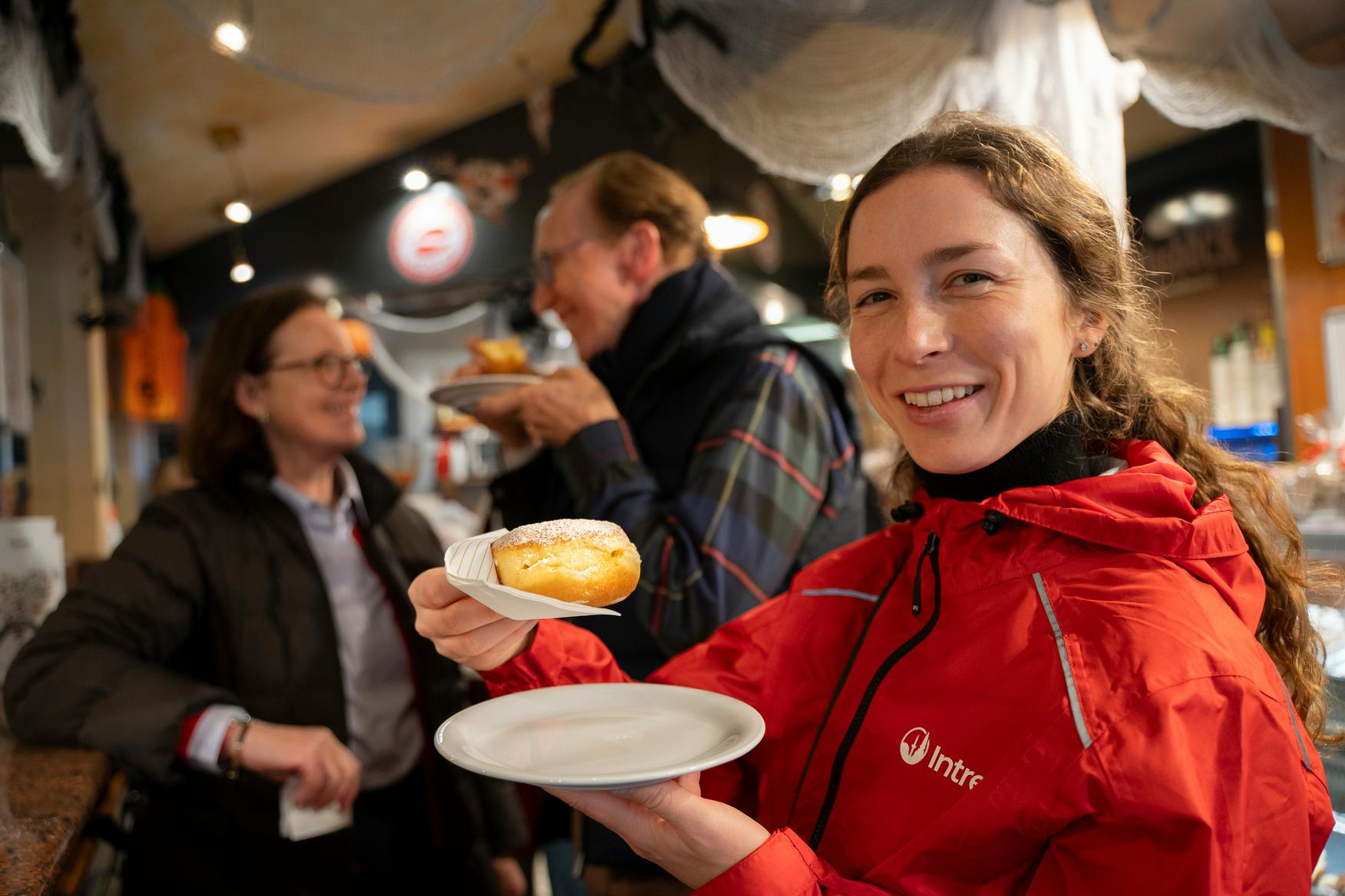 Uma pessoa de casaco vermelho sorri enquanto segura um prato com um bolo num ambiente interior, com outra pessoa em segundo plano.