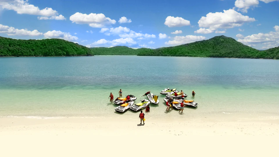People in swimsuits gather around docked jet skis on a sandy beach by a calm, clear lake surrounded by green hills.