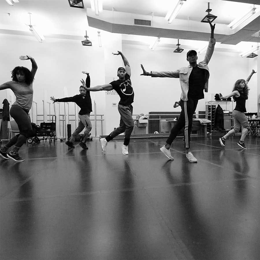 五名舞者在一个灯光充足的舞蹈工作室里摆出动态姿势，背景有镜子和设备。