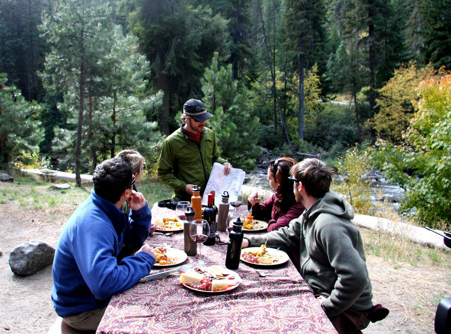 A group of people sits around a picnic table outdoors, eating, while one person stands, holding a map and talking to them.