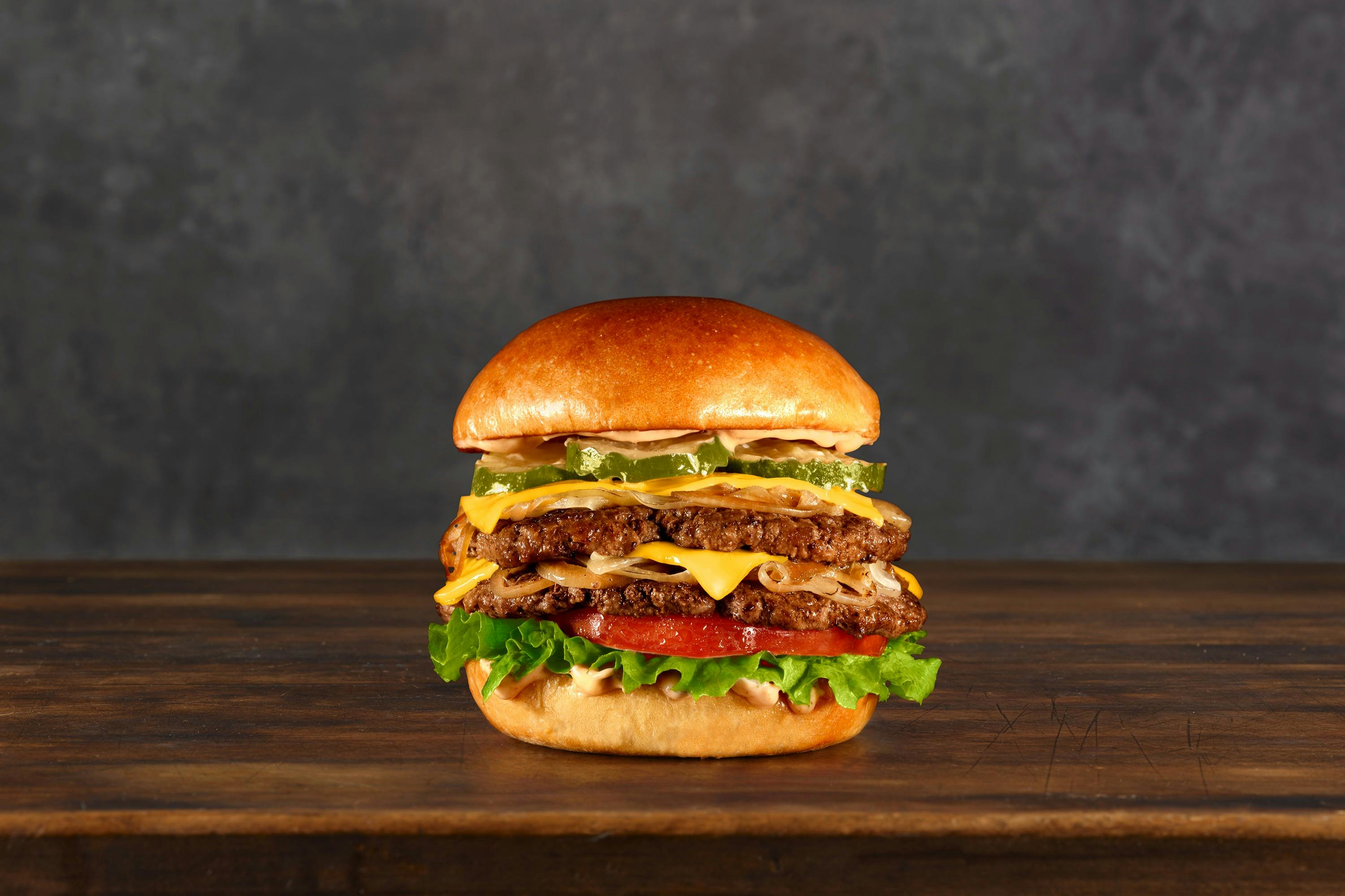 A double cheeseburger with lettuce, tomatoes, pickles, and onions is on a wooden surface with a gray background.