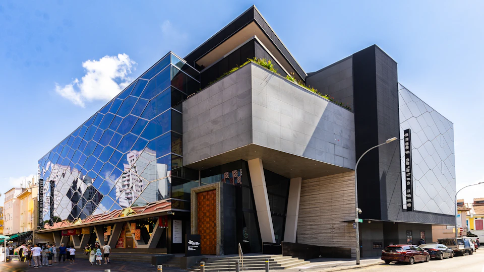 A modern building featuring a geometric glass facade, concrete walls, and a rooftop garden, under a clear blue sky.
