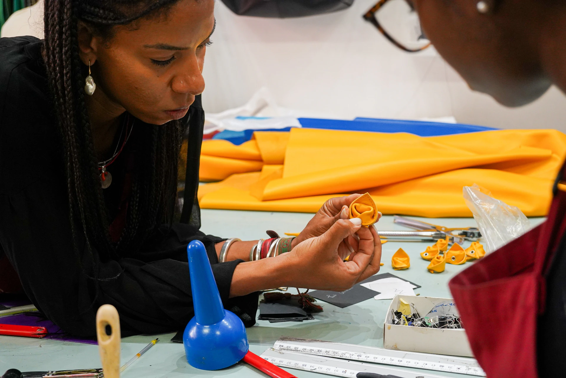 A person leaned over, viewing a small object in their hand at a craft table cluttered with tools and colored fabric.