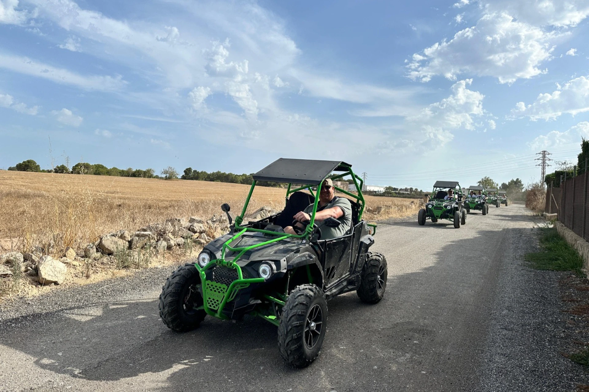 A person drives a green off-road vehicle on a rural road, with more vehicles and a dry field in the background under a partly cloudy sky.