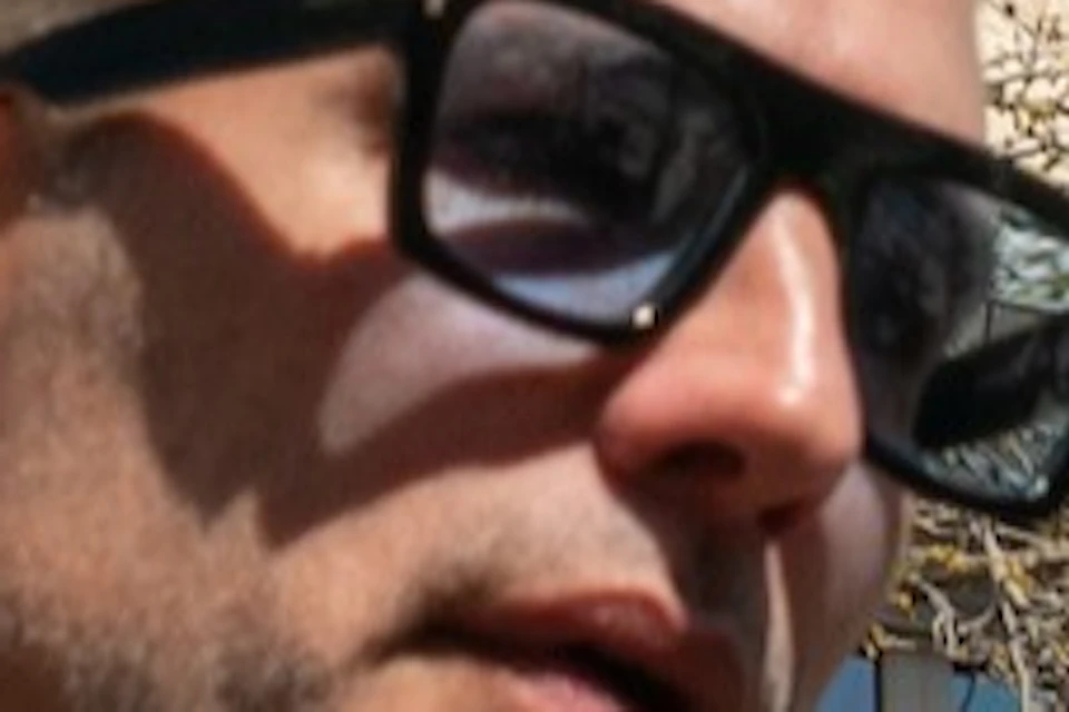 A man wearing sunglasses gestures while speaking in front of a unique building with curved balconies and intricate details.