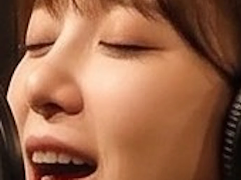 A person wearing headphones singing into a microphone in a recording studio with a music stand in the background.