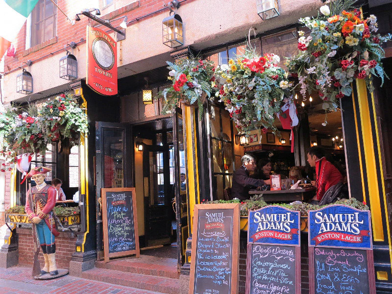 A lively pub entrance with floral decor, chalkboard menus, and patrons seated inside. Samuel Adams advertisements are prominent.