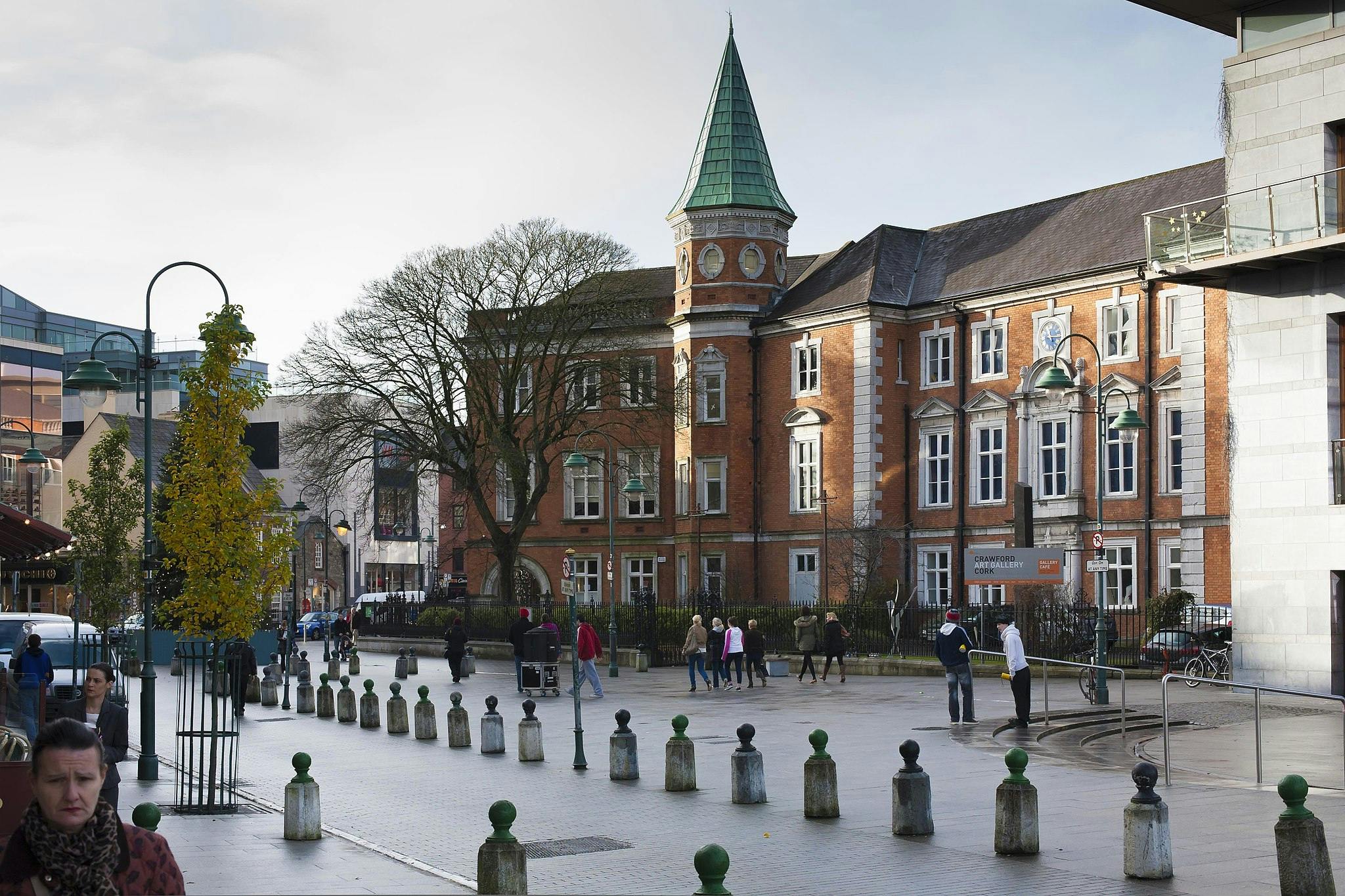 People walking near a red-brick building with a turret and a green roof, in an urban area with bollards and lampposts.
